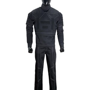 May include: Black tactical jumpsuit displayed on a mannequin. The outfit features a high collar, long sleeves, and pants, all with white stitching details. The jumpsuit has multiple panels and patches, suggesting a functional design.