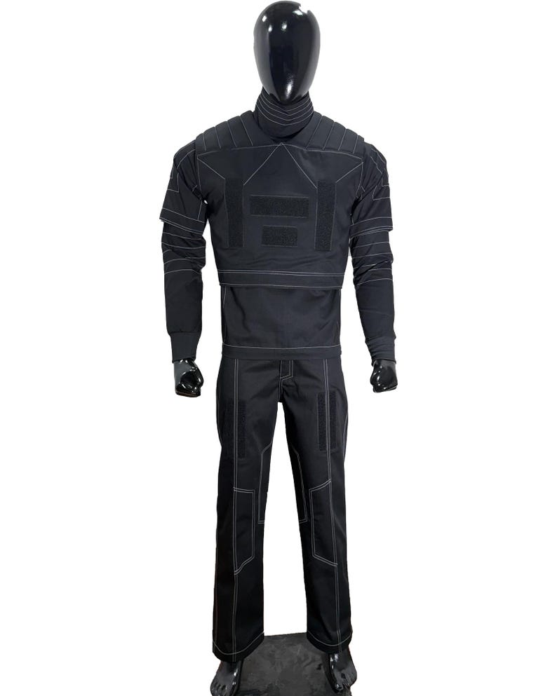May include: Black tactical jumpsuit displayed on a mannequin. The outfit features a high collar, long sleeves, and pants, all with white stitching details. The jumpsuit has multiple panels and patches, suggesting a functional design.