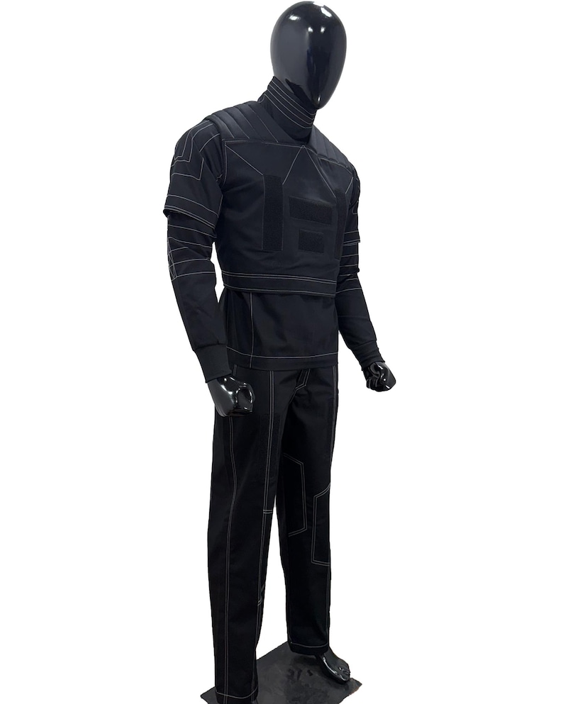 May include: A black jumpsuit with white stitching details, displayed on a mannequin. The outfit features a high collar, long sleeves, and a structured design with geometric panels. The pants are straight-legged, and the overall aesthetic is modern and sleek.