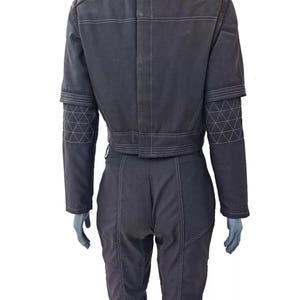 Op de afbeelding: Donkergrijze jumpsuit met witte stikseldetails. De jas heeft een korte taille, bruine schouderaccenten en geometrisch bewerkte mouwen. De broek is getailleerd met zichtbare naden. De outfit wordt op een mannequin gepresenteerd.