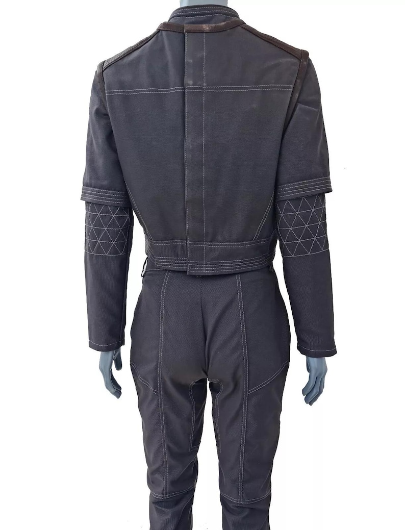 Op de afbeelding: Donkergrijze jumpsuit met witte stikseldetails. De jas heeft een korte taille, bruine schouderaccenten en geometrisch bewerkte mouwen. De broek is getailleerd met zichtbare naden. De outfit wordt op een mannequin gepresenteerd.