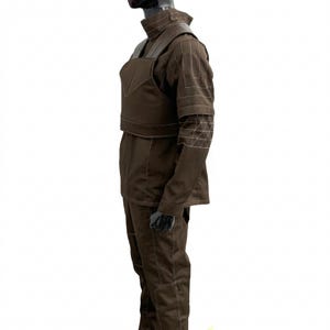 May include: A brown costume with a high collar, shoulder pads, and a chest piece. The sleeves have geometric white stitching. The trousers and jacket are brown with white stitching. The costume is displayed on a mannequin.