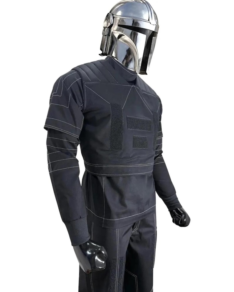 May include: A black costume with white stitching, featuring a silver helmet. The outfit includes a long-sleeved top, pants, and gloves. The top has multiple rectangular patches and a high collar. The helmet is reflective.