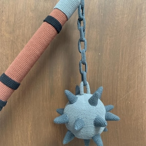 Peut inclure: Jouet de masse en crochet. Le manche est marron avec des bandes noires et orange, surmonté de gris. Une chaîne grise relie une boule à pointes grise. Le jouet est en fil doux.