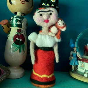 May include: A handmade felt doll resembling Frida Kahlo, wearing a black hat with white flowers, a white blouse, and a red skirt with yellow and black trim. A small orange monkey is perched on her shoulder. Other decorative items are in the background.