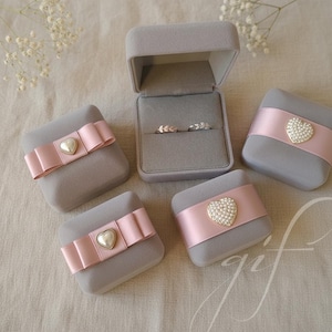 Puede incluir: Cajas grises para anillos con lazos rosas y adornos en forma de corazón. Una caja está abierta, mostrando dos anillos. Las cajas están dispuestas sobre una superficie de color neutro, ideal para bodas o compromisos.