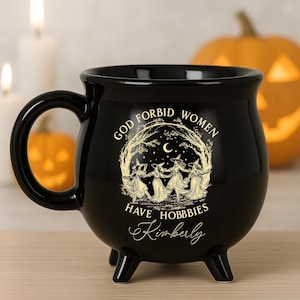 May include: Black cauldron-shaped mug with a handle and three small feet. The mug features the text "GOD FORBID WOMEN HAVE HOBBIES" with an illustration of witches dancing in a circle. The name "Kimberly" is written below the text.