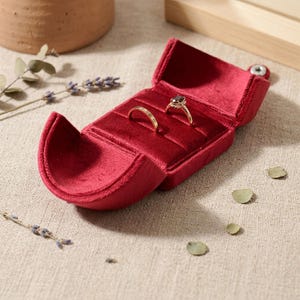 May include: A deep red velvet ring box with a hinged lid, featuring two interior slots designed to hold jewelry. Two gold-toned rings are positioned within the velvet-lined compartments, showcasing a smooth band and a ring with a dark stone.