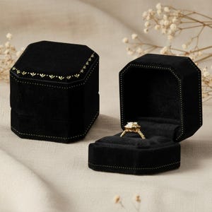Könnte beinhalten: Zwei schwarze Samt-Ringboxen, eine davon geöffnet, um einen goldenen Ring mit einem Diamanten zu zeigen. Die Boxen sind achteckig mit goldenen Verzierungen und einem dekorativen Muster auf dem Deckel. Der Ring ist auf einem schwarzen Samt-Innenraum gesetzt.