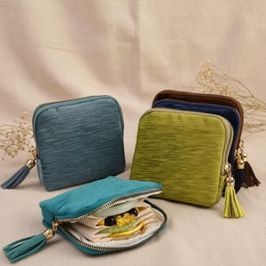 May include: A collection of small, square, zippered pouches in various colors: teal, blue, green, and brown. Each pouch has a tassel attached to the zipper pull. One pouch is open, revealing jewelry inside. The pouches are made of a textured fabric.