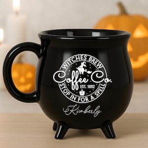 Puede incluir: Taza de café negra en forma de caldero con asa y tres patas pequeñas. La taza presenta el texto "Witches Brew Coffee Co. Stop in for a Spell" y el nombre "Kimberly" en escritura blanca. Una taza temática de Halloween.