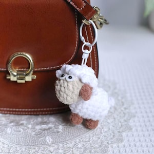 Może przedstawiać: Ręcznie robiony biały brelok do kluczy w kształcie owcy z szydełka z beżową twarzą i brązowymi uszami i kopytami. Owca jest przymocowana do białego metalowego kółka na klucze i wisi obok brązowej skórzanej torebki.