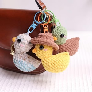 Może przedstawiać: Szydełkowe breloczki do kluczy w kształcie kaczki w kolorze żółtym, niebieskim i zielonym, z brązowym kapeluszem kowbojskim na jednym. Breloczki są przymocowane do kolorowych metalowych zapięć, wiszących w pobliżu brązowej skórzanej torebki. Kaczki mają czarne oczy i pomarańczowe dzioby.
