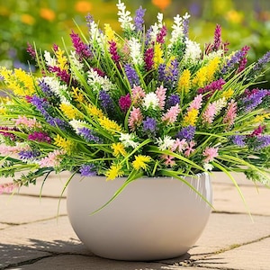 May include: A vibrant arrangement of colorful artificial flowers in a round, light gray ceramic pot. The flowers include yellow, purple, pink, and white blooms with green foliage. The pot sits on a stone surface, with a blurred background.