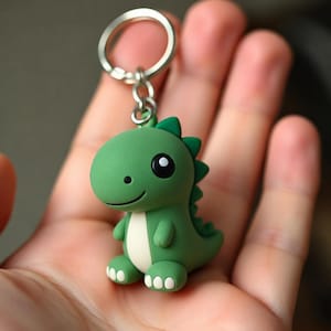 May include: A green dinosaur keychain featuring a smooth, matte finish with a cream-colored belly and white toe details. The figure has large black eyes and a small smile, attached to a silver-toned metal split ring for keys or accessories.