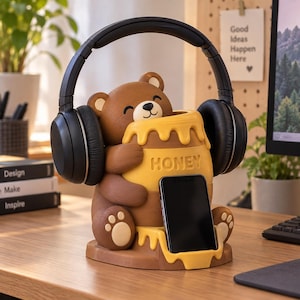 May include: A brown bear figurine holding a yellow honey pot with the word HONEY embossed on the front. The bear serves as a stand for black over-ear headphones and a black smartphone. The base features a yellow honey drip design.