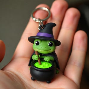 May include: A green frog keychain featuring a black witch hat with a purple band and a black cape. The frog is holding a brown stirring stick inside a black cauldron filled with bright neon green liquid, attached to a silver metal keyring.