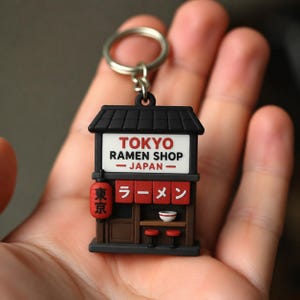 May include: A keychain featuring a miniature ramen shop building. The structure is black with a white sign reading TOKYO RAMEN SHOP JAPAN. It includes a red lantern with Japanese characters, red curtains with Japanese text, and a small bowl on a counter.