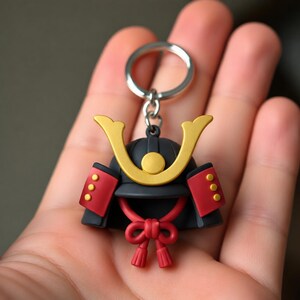 May include: A keychain featuring a miniature black samurai helmet with a gold crescent-shaped crest, red side panels with three gold dots each, and a red cord tied in a bow at the bottom. The helmet is attached to a silver metal split ring.