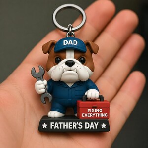 May include: A keychain featuring a brown and white bulldog figurine wearing a blue cap with the word DAD, a blue jumpsuit, and holding a grey wrench. It carries a red toolbox labeled FIXING EVERYTHING and stands on a black base marked FATHER'S DAY.