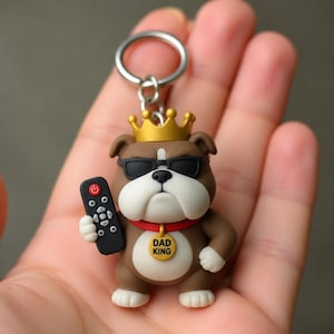 May include: A brown and white bulldog keyring featuring a gold crown, black sunglasses, and a red collar with a circular tag that reads DAD KING. The bulldog is holding a black television remote control with a red power button in its right hand.