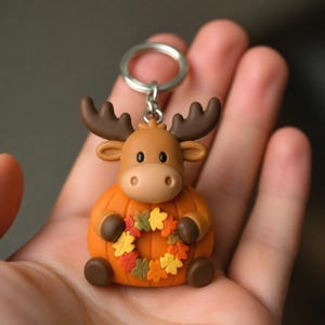 May include: A keychain featuring a brown moose character with dark brown antlers, sitting on a round orange pumpkin body. The moose holds a small wreath made of multicolored autumn leaves in its front paws, attached to a silver metal ring.