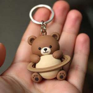 May include: A brown bear keyring featuring a cartoon bear with a cream-coloured belly and a brown ring encircling its midsection like a planet. The bear has small ears, black eyes, a black nose, and paw pads on its feet, attached to a silver ring.