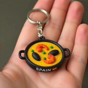 May include: A round keychain featuring a miniature black paella pan filled with yellow rice, two orange shrimp, two black mussels, and green peas. The side of the pan is printed with the word SPAIN and a small Spanish flag icon in white text.