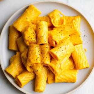 Könnte beinhalten: Draufsicht auf einen Teller mit Rigatoni-Nudeln, bedeckt mit einer cremigen, leuchtend orangefarbenen Käsesauce. Die Nudeln sind auf einem Haufen angeordnet, mit sichtbarem schwarzem Pfeffer bestreut. Der Teller ist hellgrau.