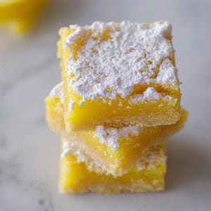 May include: A stack of three lemon bars dusted with powdered sugar. The bars have a bright yellow filling on a shortbread crust. The dessert is stacked on top of each other, with the top bar slightly angled.