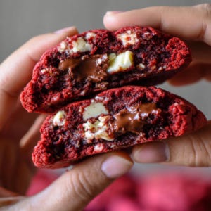 May include: A close-up of a red velvet cookie cut in half, revealing a dark chocolate center and white chocolate chunks. The cookie is held in a person's hands, showcasing its soft texture and rich color.