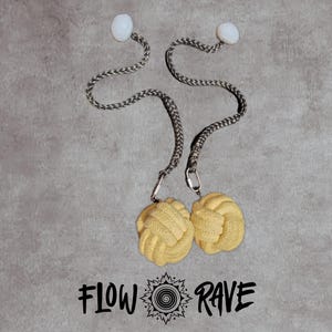 May include: Two yellow monkey fist knots attached to grey braided cords with white spherical beads at the ends and metal clips. The text FLOW RAVE with a central spiral icon is printed below the knots on the grey background surface.