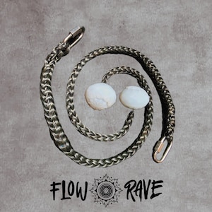 Peut inclure: Un cordon tressé gris foncé disposé en spirale avec deux extrémités sphériques blanches et des mousquetons métalliques aux bouts. Le texte FLOW RAVE avec une icône en spirale centrale est imprimé sous le cordon sur un fond gris texturé.