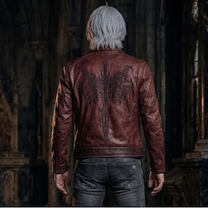 May include: A burgundy leather jacket with a black graphic design on the back. The jacket has a classic, fitted style with a front zipper and a small collar. The design on the back features a sword with wing-like details.