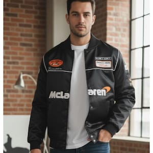 Può includere: Giacca bomber in raso nero con profili bianchi e toppe multiple. La giacca presenta il logo McLaren in bianco e arancione. La giacca ha una chiusura con bottoni e colletto e polsini a coste. La giacca è indossata sopra una maglietta bianca.