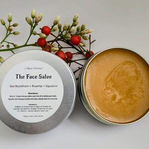 May include: A silver tin of face salve with the text "The Face Salve" and ingredients listed. The open tin reveals a golden-brown balm. The product is 1 oz / 40 ml. A sprig of berries and leaves is in the background.