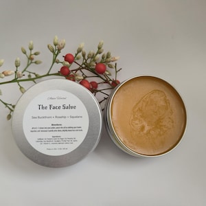 May include: Two open silver tins of face salve with a light brown, creamy texture. The lid of one tin displays the text "The Face Salve" and ingredients. A sprig of white flowers and red berries is in the background.