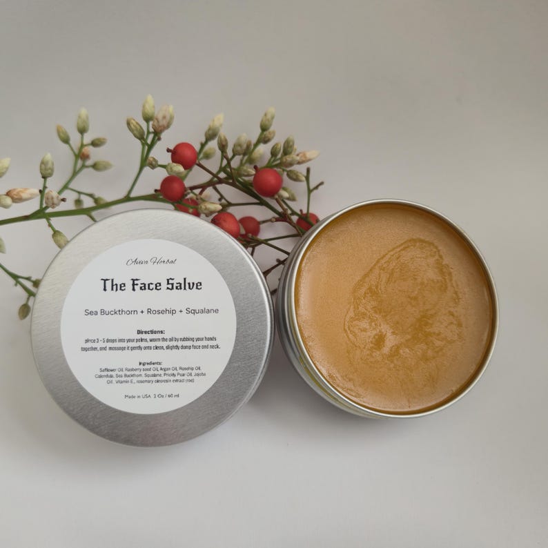 May include: Two open silver tins of face salve with a light brown, creamy texture. The lid of one tin displays the text "The Face Salve" and ingredients. A sprig of white flowers and red berries is in the background.