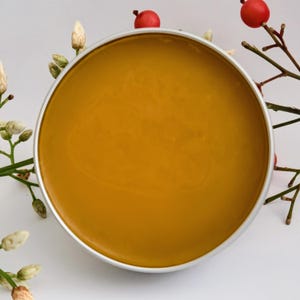 May include: A round, silver-rimmed container filled with a smooth, golden-yellow substance. The container is surrounded by sprigs of white flowers and branches with red berries, set against a white background. The product appears to be a cosmetic or skincare item.
