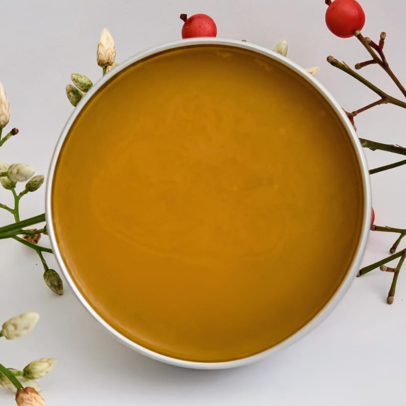 May include: A round, silver-rimmed container filled with a smooth, golden-yellow substance. The container is surrounded by sprigs of white flowers and branches with red berries, set against a white background. The product appears to be a cosmetic or skincare item.