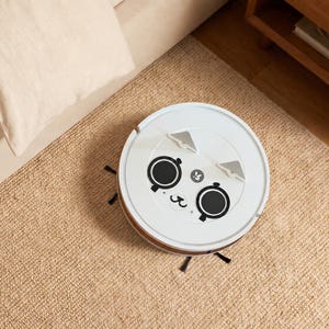 May include: A white circular robotic vacuum cleaner featuring a decorative face decal with two large black circular eyes, a small mouth, and two triangular grey ears positioned on the top surface. The device has black side brushes extending outwards.