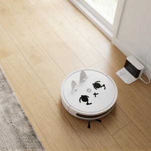 Puede incluir: Una aspiradora robótica blanca con un diseño de cara de dibujos animados, que incluye orejas y rasgos faciales. La aspiradora está en un piso de madera clara cerca de una estación de carga y una alfombra.