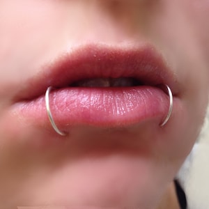 May include: Close-up of a person's lower lip with two silver lip rings. The rings are thin and circular, and they are placed on either side of the lip.