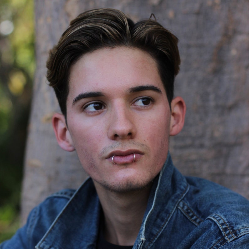May include: A young person with short brown hair wearing a blue denim jacket. They have a silver lip ring.