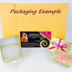 May include: A gold-colored box with a white interior and a small gold ring inside. The box is sitting on a white surface. A business card with the text "Dazzling Adornments Quality Handmade Cuffs" is visible in the background. The business card also includes contact information for the company.