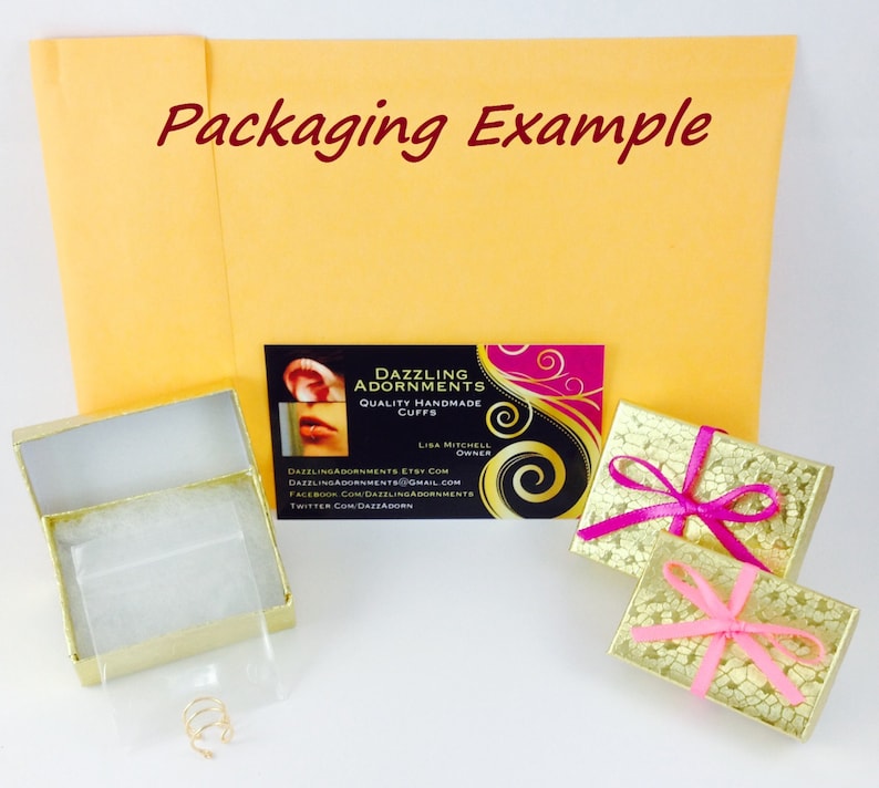 May include: A gold-colored box with a white interior and a small gold ring inside. The box is sitting on a white surface. A business card with the text "Dazzling Adornments Quality Handmade Cuffs" is visible in the background. The business card also includes contact information for the company.