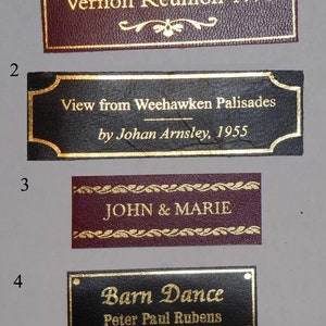 May include: Four rectangular leather plaques with gold trim and lettering. The top plaque is maroon with gold lettering that reads "Vernon Reunion 1998". The second plaque is black with gold lettering that reads "View from Weehawken Palisades by Johan Arnsley, 1955". The third plaque is maroon with gold lettering that reads "JOHN & MARIE". The bottom plaque is black with gold lettering that reads "Barn Dance Peter Paul Rubens".