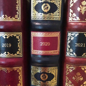 Może przedstawiać: Trzy czerwone książki w skórzanych oprawach z złotymi zdobieniami i czarnymi grzbietami. Książki są oznaczone datami 2019, 2020 i 2021.