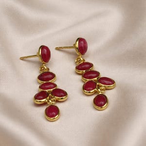 May include: A pair of gold-colored dangle earrings with a cluster of oval, deep red gemstones. The earrings have a post-back closure and are set against a soft, cream-colored fabric.