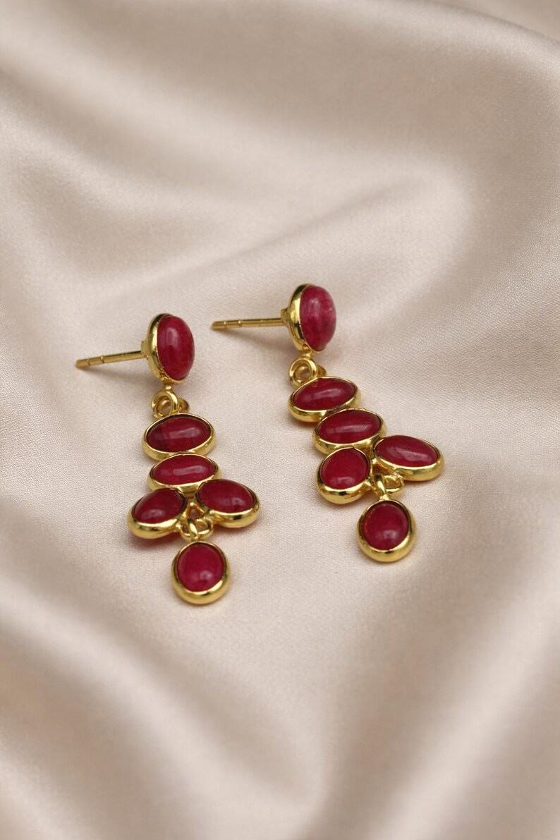 May include: A pair of gold-colored dangle earrings with a cluster of oval, deep red gemstones. The earrings have a post-back closure and are set against a soft, cream-colored fabric.